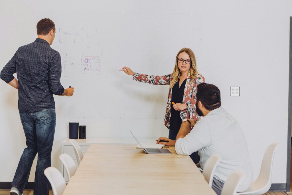 Photo by Jud Mackrill man standing and facing whiteboard with writings near woman pointing at it and facing man sitting beside table with laptop indoors